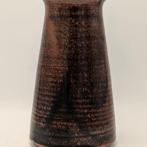 May include: A tapered ceramic vase with a flared rim, featuring a textured surface with a mottled finish in shades of dark brown and black. The vessel has a wide base that narrows towards the top, showcasing a rustic, earthy appearance throughout.