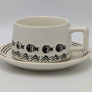 May include: Cream ceramic cup and saucer set. The cup has a square handle and is decorated with black fish and wave patterns. The saucer has a matching design. A charming addition to any kitchen or dining room.