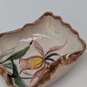 May include: A white ceramic dish with a brown rim and a floral design. The dish has a scalloped edge and features a pink and yellow flower with green leaves.