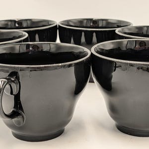 May include: Six black teacups with curved handles are arranged together. The cups have a glossy finish and a simple, elegant design. The cups are likely made of ceramic or porcelain and are suitable for serving tea or coffee.
