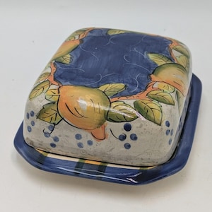 May include: A ceramic butter dish with a rectangular base and lid. The lid features a blue background and a pattern of yellow lemons and green leaves. The base is blue with a plaid design.