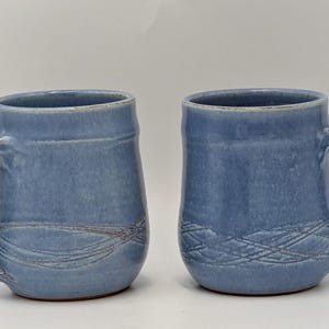May include: Two blue ceramic mugs with curved handles. Each mug has a textured surface and a decorative etched design near the base. The mugs are displayed against a plain white background.