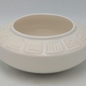 May include: A cream-coloured, low, round ceramic bowl. The bowl has a wide, flat base and a slightly raised rim. The exterior features a repeating geometric pattern. The interior is a lighter cream colour.