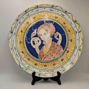 May include: A decorative ceramic plate with a portrait of a person, set within a blue and yellow floral pattern. The portrait is on a blue background, with the name "Bartolo" visible. The plate is supported by a black stand.
