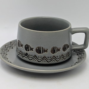 May include: A grey ceramic teacup and saucer set. The cup has a handle and is decorated with a repeating pattern of fish and wavy lines. The saucer has a matching design. The set is likely vintage.