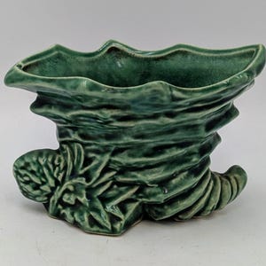 May include: A green ceramic vase with a unique, textured design. The vase has a wavy rim and a swirling, organic shape, with a leaf-like detail on the side. The vase is approximately 15 cm tall.