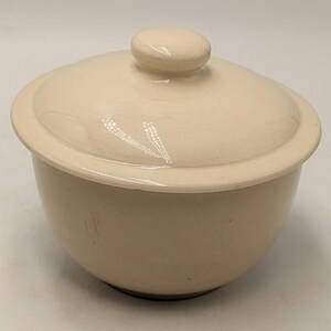 May include: A cream-coloured ceramic bowl with a matching lid featuring a rounded knob handle. The bowl has a smooth, glossy finish with subtle speckling throughout the surface, a wide rim, and a deep, rounded body shape for holding contents.