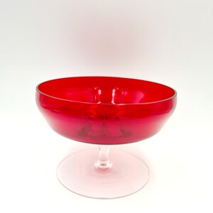 May include: A red glass bowl with a clear glass pedestal base. The bowl is round and has a wide rim.