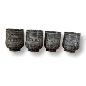 May include: Set of four small, brown ceramic cups with white floral designs. The cups have a rounded shape and a slightly flared lip.