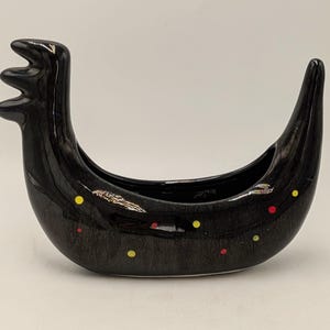 May include: A black ceramic decorative vessel in a bird-like form. The vessel has a curved body, a tall, three-pronged head, and a pointed tail. It is decorated with small, colourful dots in yellow and red.