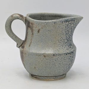 May include: A handmade ceramic jug with a speckled, textured surface. The jug is primarily light blue-grey with darker grey and brown accents. It features a rounded body, a spout for pouring, and a curved handle.