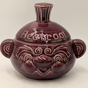 May include: A dark purple ceramic lidded jar with two side handles and a knobbed lid. The word BEETROOT is embossed on the lid. The body features a raised, swirling relief pattern. The finish is glossy and uniform in colour throughout the piece.