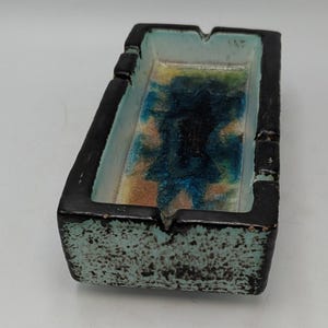 May include: A rectangular ceramic ashtray featuring a thick black outer rim with a textured light blue speckled finish. The interior basin displays a vibrant glaze of blended blue, green, and yellow tones, with four notches along the edges.