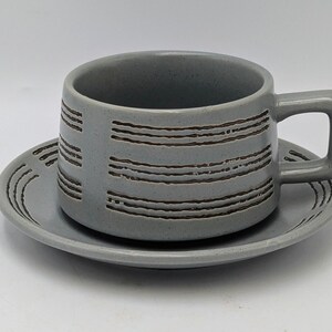 May include: A grey ceramic teacup and saucer set. The cup has a square handle and is decorated with horizontal brown lines. The saucer has matching brown lines. The set is designed for serving tea or coffee.