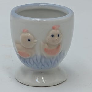 May include: White ceramic egg cup featuring two baby chicks on the front. The chicks are pink and yellow with blue eyes. The egg cup has a blue and white design around the base.