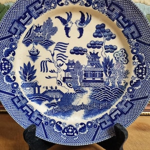 May include: A blue and white porcelain plate with a traditional Chinese design. The plate features a pagoda, a willow tree, and two birds in flight. The border of the plate is decorated with a repeating pattern of blue and white.