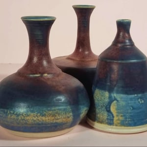 May include: Three handcrafted ceramic vases in shades of blue and brown. The vases have unique shapes, including a bulbous form, a tall slender neck, and a bell-shaped design. The glaze creates a textured, artistic appearance, with hints of yellow and green.