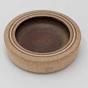 May include: A circular, beige ceramic ashtray with a dark brown interior. Two concentric brown lines encircle the rim. The ashtray has a textured surface and a small, circular mark on the side.