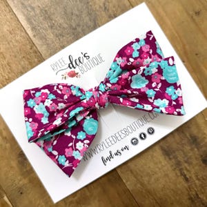 May include: A large pink and teal floral print bow. The bow is tied with a knot in the center. The bow is on a white card with the text "Rylee dee's Boutique" and "www.ryleedeesboutique.com find us on" with social media icons.