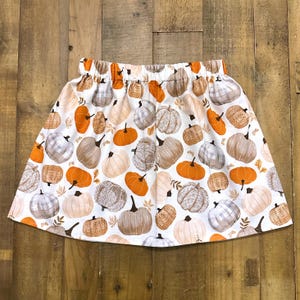 Puede incluir: Falda blanca con cintura elástica, estampada con varias calabazas en tonos naranja, marrón y gris. La falda está hecha de una tela ligera, perfecta para el otoño. La falda está diseñada para niños.