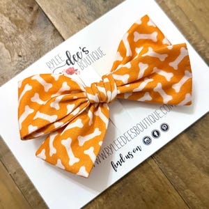 May include: An orange and white dog bone patterned hair bow. The bow is tied in the center and is on a white card with the text "Rylee Dee's Boutique" and social media icons.