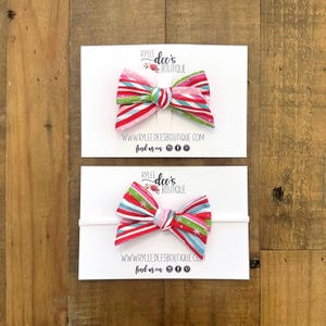 May include: Two fabric bows with red, pink, green, and blue stripes and a white background. One bow is attached to a white headband. The bows are on white cards with the text "RYLEE dee's BOUTIQUE" and a website address.