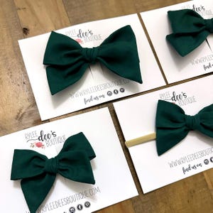 May include: Several dark green fabric bows on white cards. Each bow is centered on the card. The cards have the text "RYLEE dee's BOUTIQUE" and "WWW.RYLEEDEESBOUTIQUE.COM" printed on them. Some bows are attached to headbands.