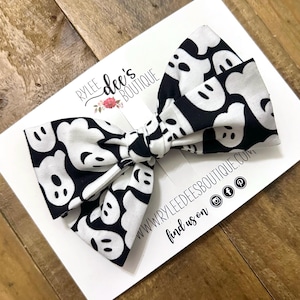 May include: A black and white patterned hair bow featuring a ghost design. The bow is on a white card with the text "Rylee Dee's Boutique" and social media icons. The bow is tied in the center.
