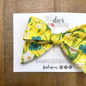 May include: A yellow floral hair bow with white and teal flowers. The bow is on a white card with the text "RYLEE dee's BOUTIQUE" and social media icons. The bow is made of fabric and is tied in the center.