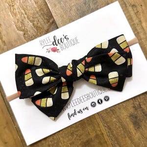 May include: A black bow with a candy corn pattern, set on a nude headband. The bow features a repeating design of orange, yellow, and white candy corn shapes. The product is displayed on a white card with the text "Rylee dee's boutique".