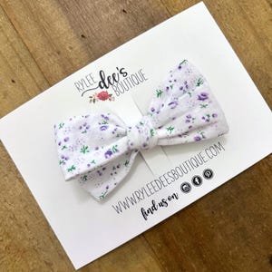 May include: White fabric bow with a purple and green floral pattern. The bow is on a white card with the text "Rylee dee's Boutique" and "www.ryleedeesboutique.com find us on" with social media icons.