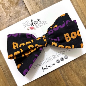 May include: A black fabric bow with the word "Boo" in orange and purple letters. The bow is on a white card with the text "Rylee Dee's Boutique" and social media icons.