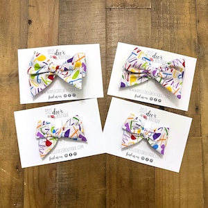 May include: Four white fabric hair bows with colorful letter and shape patterns. Each bow is on a white card with the text "RYLEE dee's BOUTIQUE" and "www.RYLEEDEESBOUTIQUE.COM". The bows are arranged on a wooden surface.