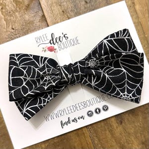 May include: A black bow tie with a silver spiderweb pattern. The bow tie is on a white card with the text "Rylee Dee's Boutique" and social media icons. The bow tie is likely for Halloween or a themed event.