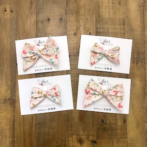 May include: Four pink fabric hair bows with floral patterns, each displayed on a white card. The bows have pink, green, and blue floral designs. The cards have the text "RYLEE dee's BOUTIQUE" and a website address.