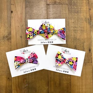 May include: Three floral fabric hair bows on white cards. The bows feature a colorful pattern of yellow, pink, purple, and blue flowers. Each card has the text "RYLEE dee's BOUTIQUE" and a website address.