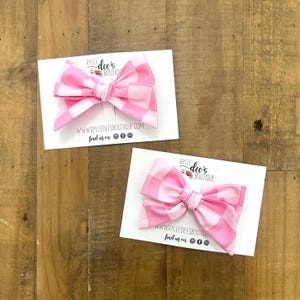 May include: Two pink and white checkered fabric bows on white cards. The bows are tied in the center and have long, flowing ends. The cards have the text "RYLEE dee's BOUTIQUE" and "WWW.RYLEEDEESBOUTIQUE.COM find us on" printed on them.