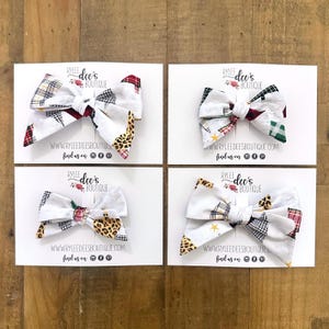 May include: Four handmade fabric hair bows with various patterns, including plaid, leopard print, and stars, are displayed on white cards. Each bow is unique, with a mix of colors and textures. The cards feature the text "RYLEE dee's BOUTIQUE" and a website address.