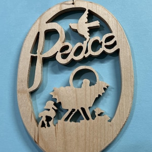 May include: Wooden Christmas ornament with the word "Peace" cut out in an oval shape. A dove and a nativity scene are also cut out within the oval. The ornament is made of light-colored wood and has a gold string for hanging.