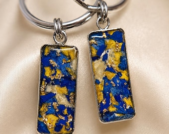 May include: Two rectangular keychains featuring a silver-toned metal frame and a resin inlay. The inlay contains a mosaic of blue and yellow fragments accented with small flecks of gold leaf. Each pendant is attached to a circular silver metal ring.