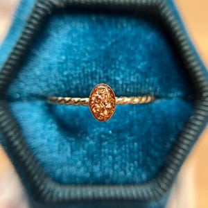 May include: A gold-toned ring featuring a twisted band and a central oval-shaped setting. The oval centerpiece contains a textured, amber-colored resin inlay with embedded light-colored flecks that create a shimmering, multi-dimensional appearance.