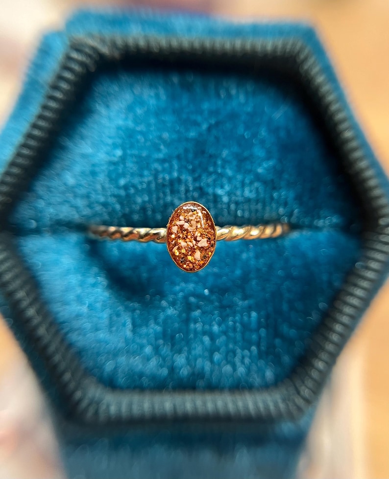 May include: A gold-toned ring featuring a twisted band and a central oval-shaped setting. The oval centerpiece contains a textured, amber-colored resin inlay with embedded light-colored flecks that create a shimmering, multi-dimensional appearance.