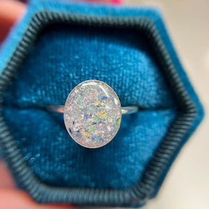 May include: A silver ring featuring an oval-shaped setting filled with a translucent resin containing fine, multi-colored glitter particles in shades of blue, orange, green, and white, creating a speckled, light-reflecting appearance.