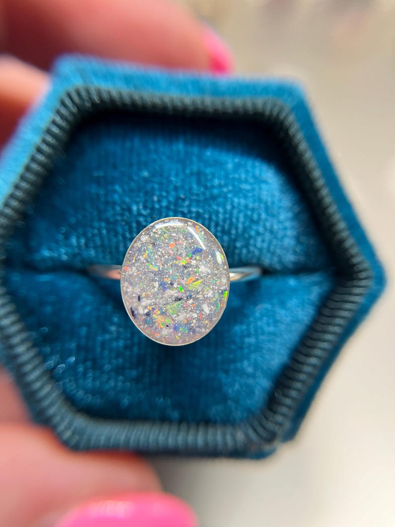 May include: A silver ring featuring an oval-shaped setting filled with a translucent resin containing fine, multi-colored glitter particles in shades of blue, orange, green, and white, creating a speckled, light-reflecting appearance.