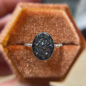 May include: A silver ring featuring an oval-shaped setting filled with a dark, speckled inlay containing small, multicolored flecks of red, green, and white. The band of the ring has a twisted, rope-like texture, providing a distinct metallic finish.