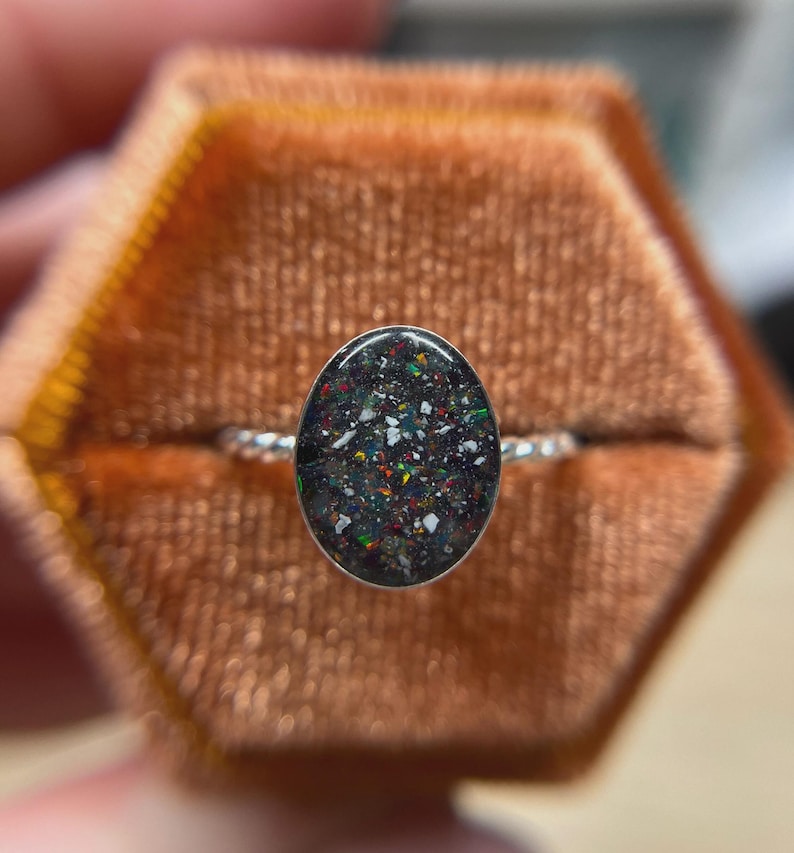 May include: A silver ring featuring an oval-shaped setting filled with a dark, speckled inlay containing small, multicolored flecks of red, green, and white. The band of the ring has a twisted, rope-like texture, providing a distinct metallic finish.