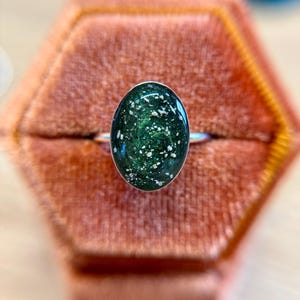 May include: A silver ring featuring an oval-shaped cabochon with a deep forest green, swirling resin base embedded with fine metallic glitter and small, irregular white flecks that resemble crushed stone or mineral inclusions.