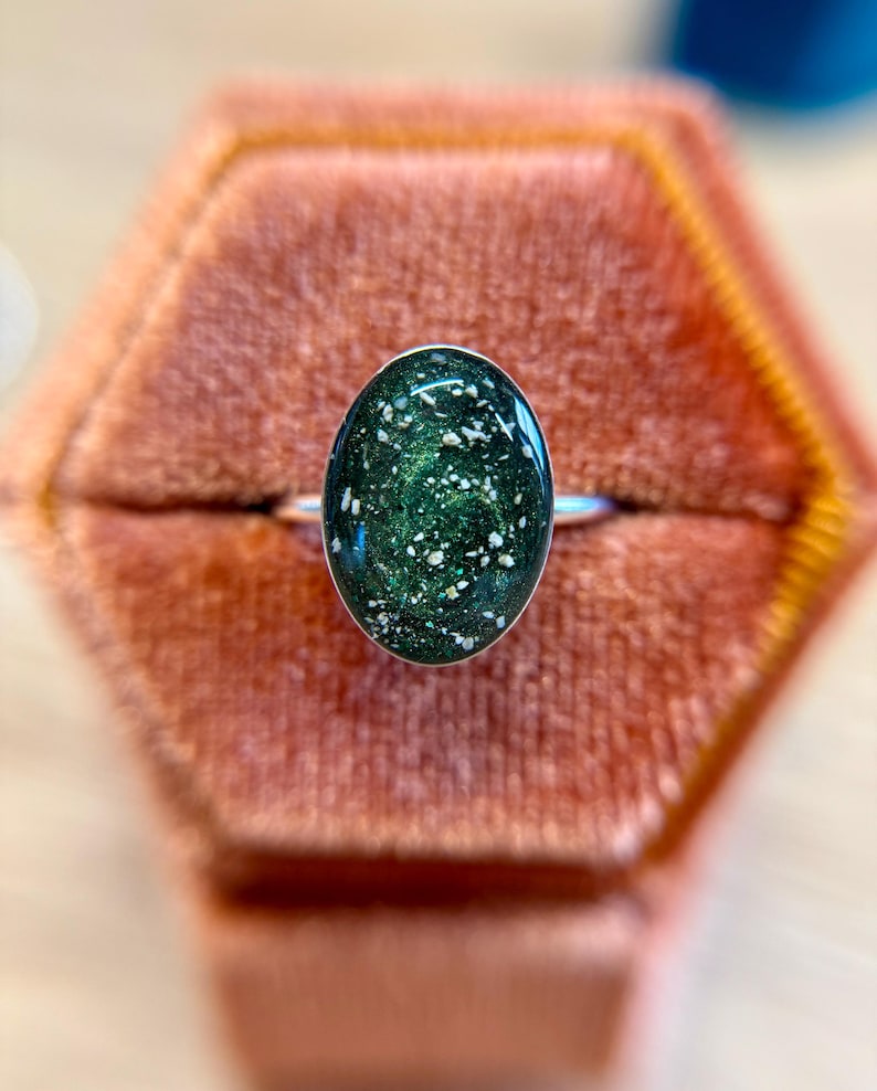May include: A silver ring featuring an oval-shaped cabochon with a deep forest green, swirling resin base embedded with fine metallic glitter and small, irregular white flecks that resemble crushed stone or mineral inclusions.