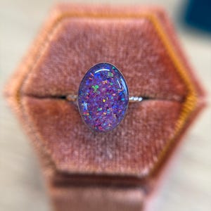 May include: A ring featuring an oval-shaped cabochon with a purple base and embedded iridescent flecks in shades of blue, green, and pink. The stone is set in a metallic frame with a thin, twisted band, creating a reflective and textured appearance.