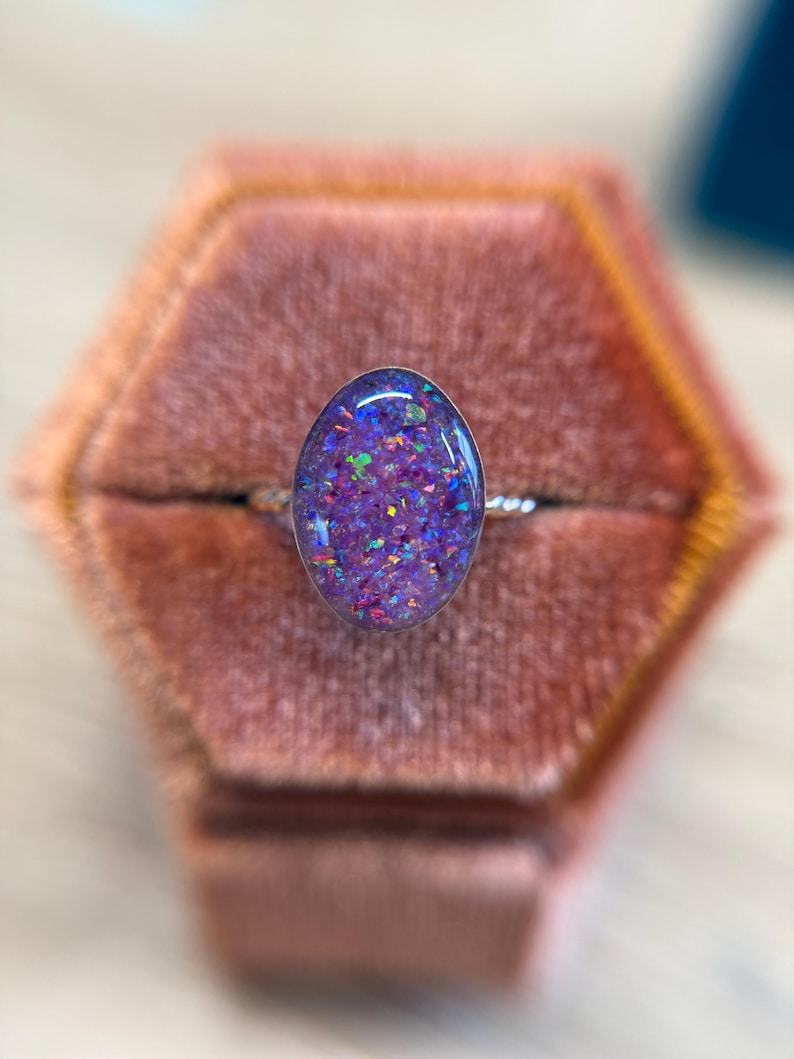 May include: A ring featuring an oval-shaped cabochon with a purple base and embedded iridescent flecks in shades of blue, green, and pink. The stone is set in a metallic frame with a thin, twisted band, creating a reflective and textured appearance.